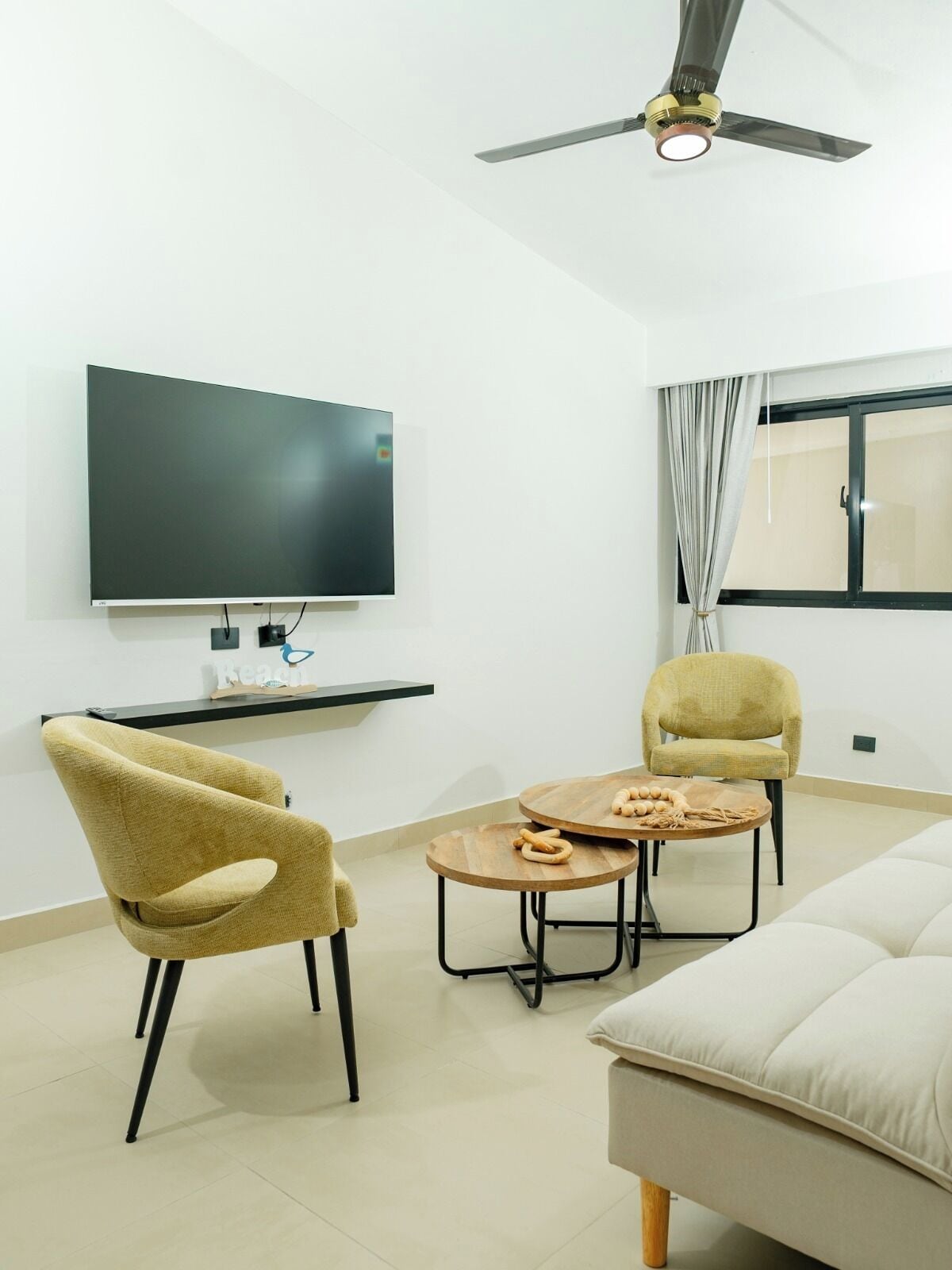 A modern living room with a flat-screen TV, two chairs, a round wooden table, a ceiling fan, and a large window with curtains.
