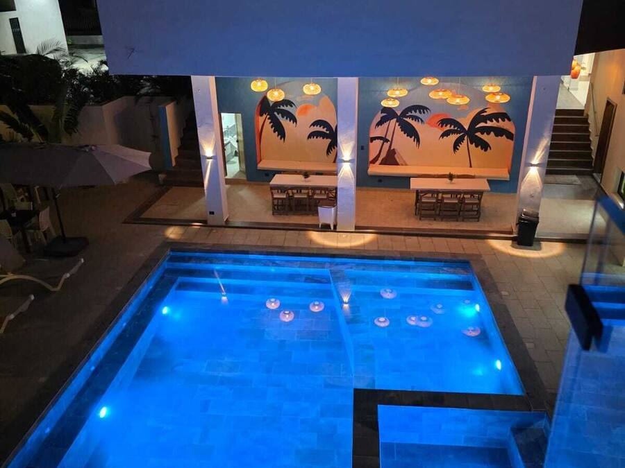 A swimming pool with a blue-lit design, a seating area with tables and chairs, and a mural of palm trees on the wall.
