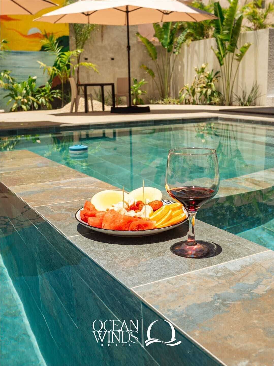 A poolside setting with a plate of fruit and a glass of red wine, a lounge chair, and an umbrella.