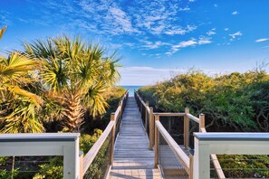 Ein hölzerner Steg führt zu einem Strand, umgeben von üppiger Vegetation und Palmen.