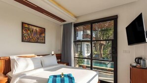 A hotel room with a large bed, a painting on the wall, a TV mounted on the wall, and a window with a view of a pool and greenery. - Paripadi Studio Canggu (Bali)