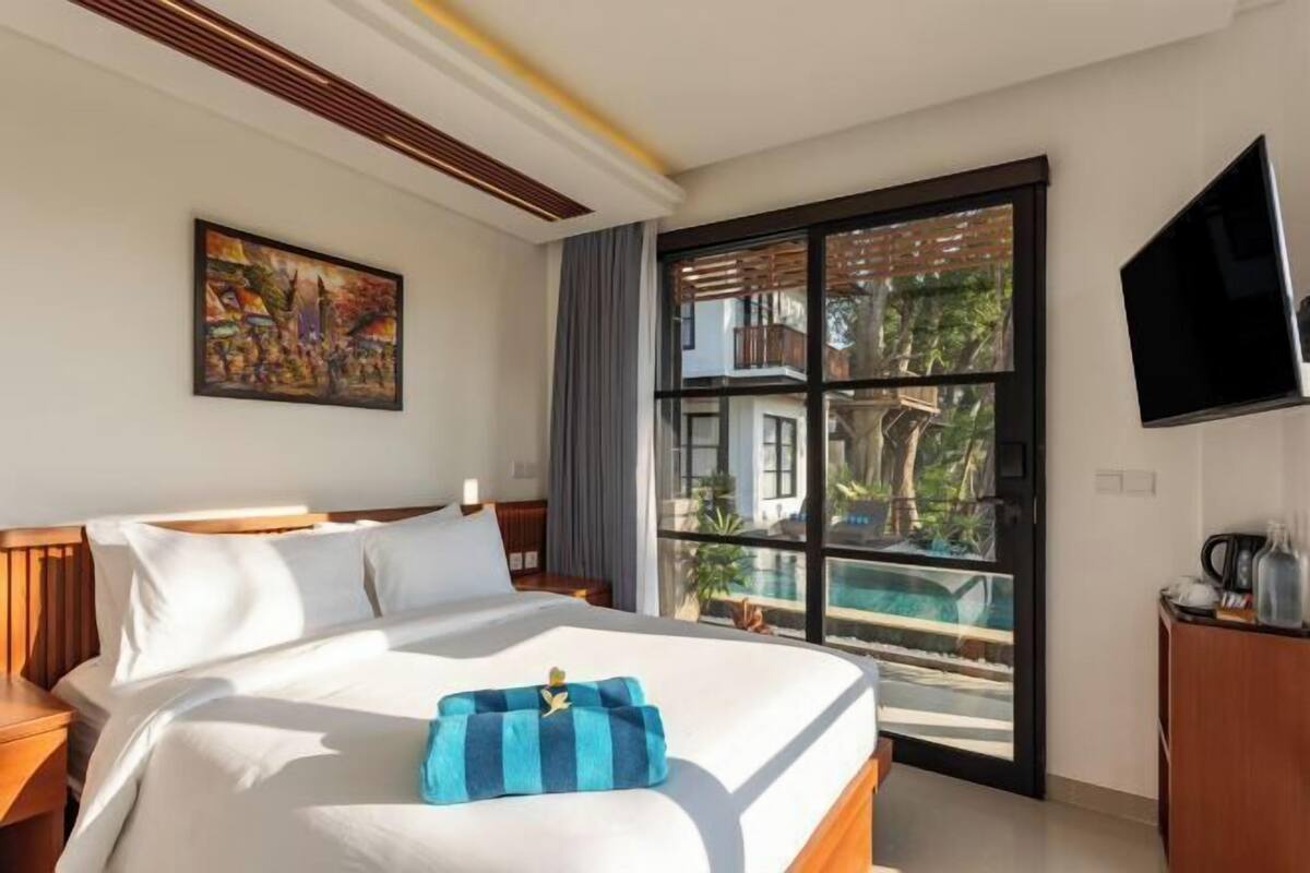 A hotel room with a large bed, a painting on the wall, a TV mounted on the wall, and a window with a view of a pool and greenery.