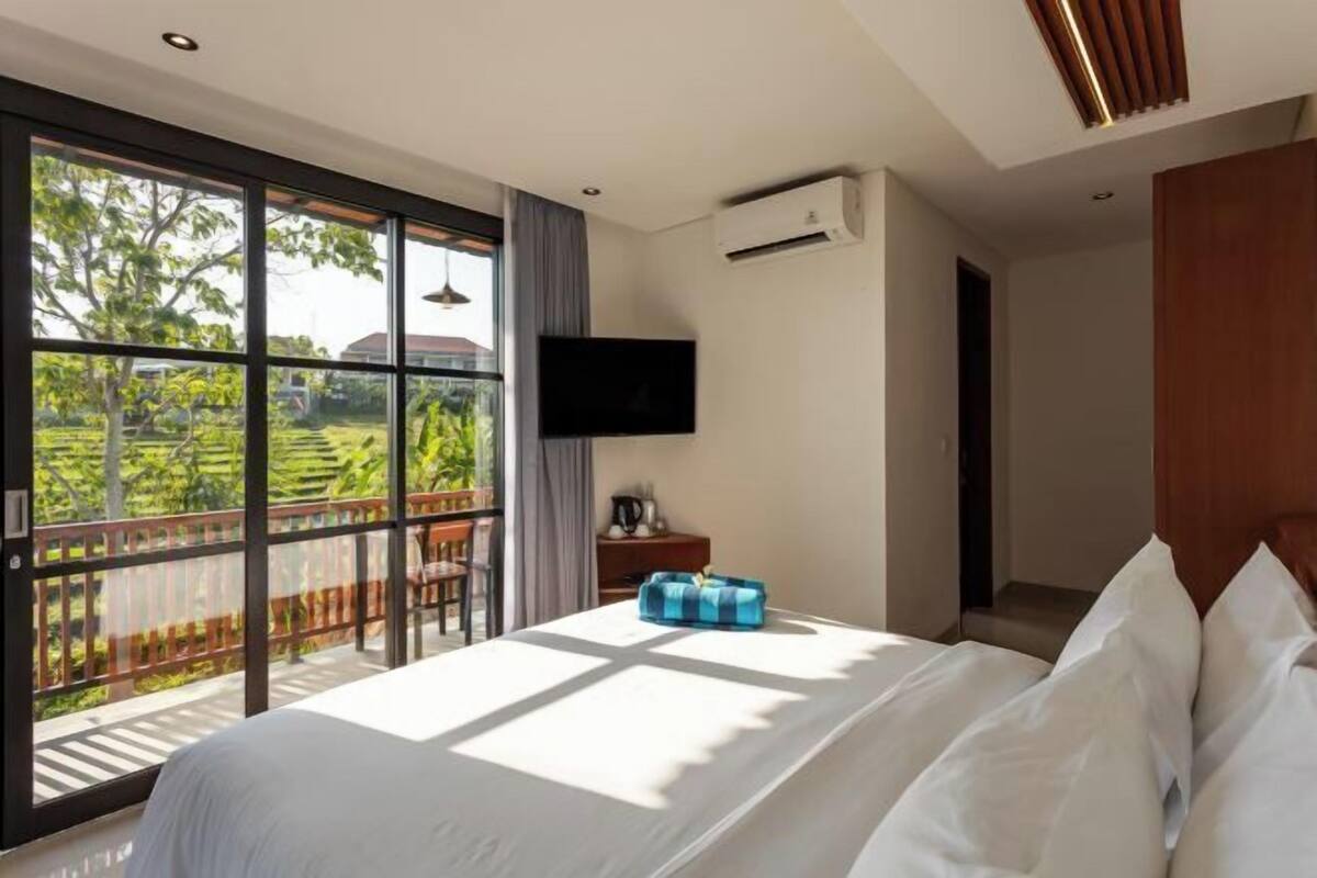 A hotel room with a large bed, a TV mounted on the wall, and a balcony with a view of greenery.
