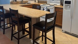 A kitchen with wooden cabinets, a white refrigerator, a stove, and a granite countertop. There are four dark wooden chairs around the counter.