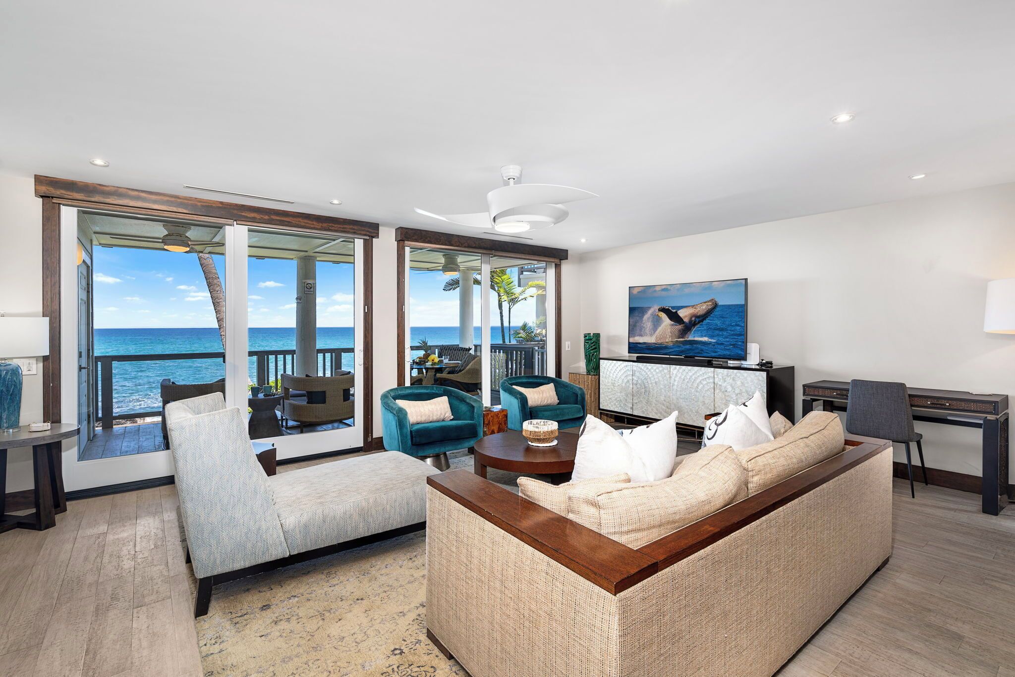 A modern living room with a large sofa, two armchairs, and a coffee table. There is a television and a desk with a lamp. The room has large windows with a view of the ocean.