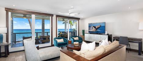 A modern living room with a large sofa, two armchairs, and a coffee table. There is a television and a desk with a lamp. The room has large windows with a view of the ocean.