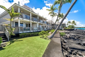A two-story beachfront property with a balcony, palm trees, and a rocky shoreline. - Luxury Oceanfront Panoramic Views, Private Sauna & Beach Access - Vrkcv (Kailua-Kona)