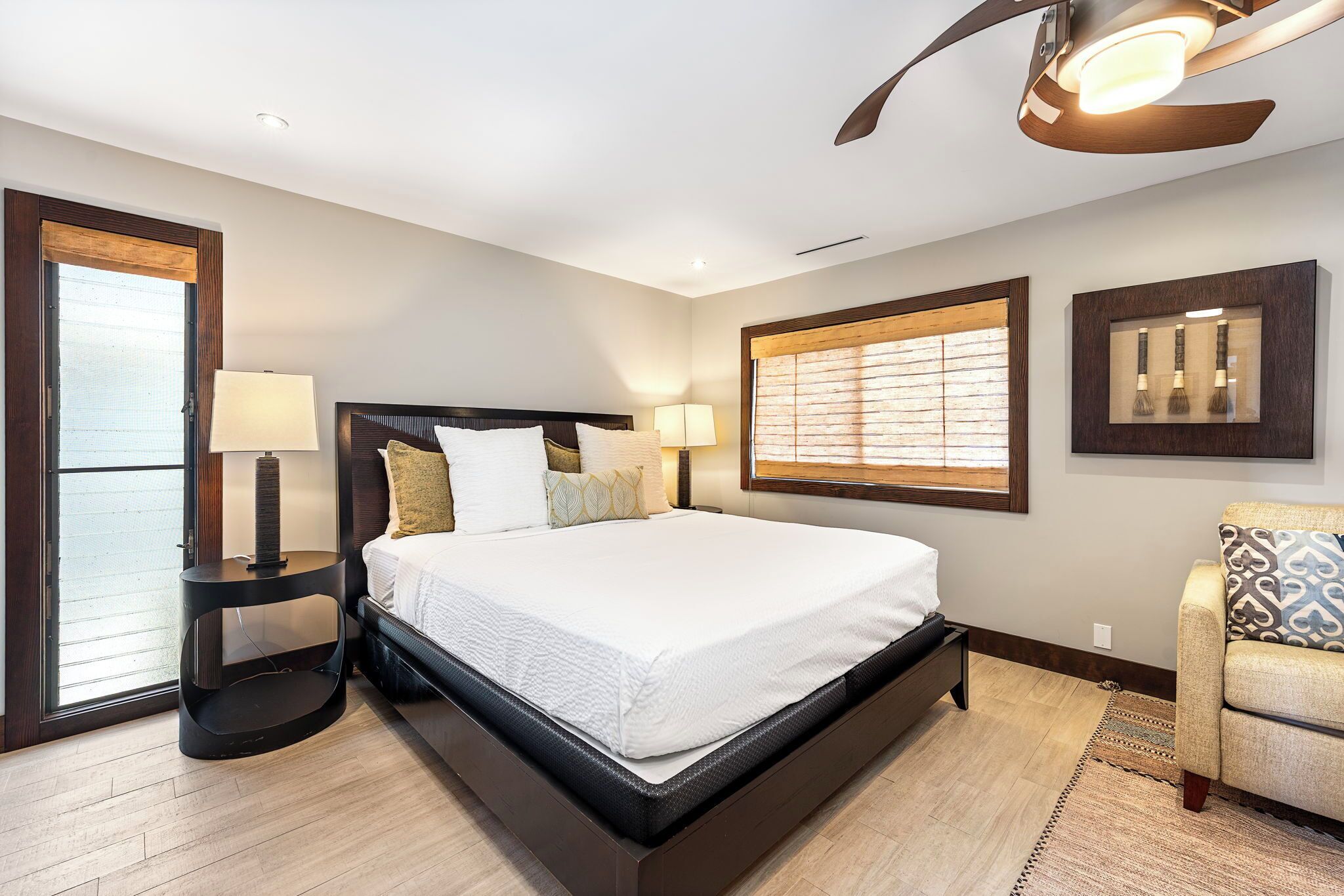 A modern bedroom with a large bed, bedside lamps, a ceiling fan, and a view through a window with blinds.