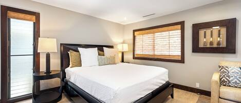 A modern bedroom with a large bed, bedside lamps, a ceiling fan, and a view through a window with blinds.