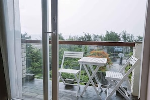 Un balcon avec une table et deux chaises, une porte vitrée, et une vue sur la verdure et une voiture garée.