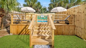 Une terrasse en bois, agrémentée d’une pancarte indiquant « Bienvenue à la plage », mène à une zone de piscine, entourée de palmiers et de parasols.