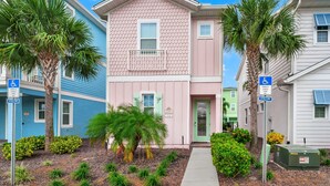 A row of colorful townhouses with a central pink building, each with a wheelchair-accessible sign, a sidewalk, and a small garden area.