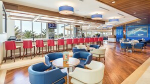 A modern restaurant with wooden ceilings, blue and red chairs, and large windows offering views of palm trees.