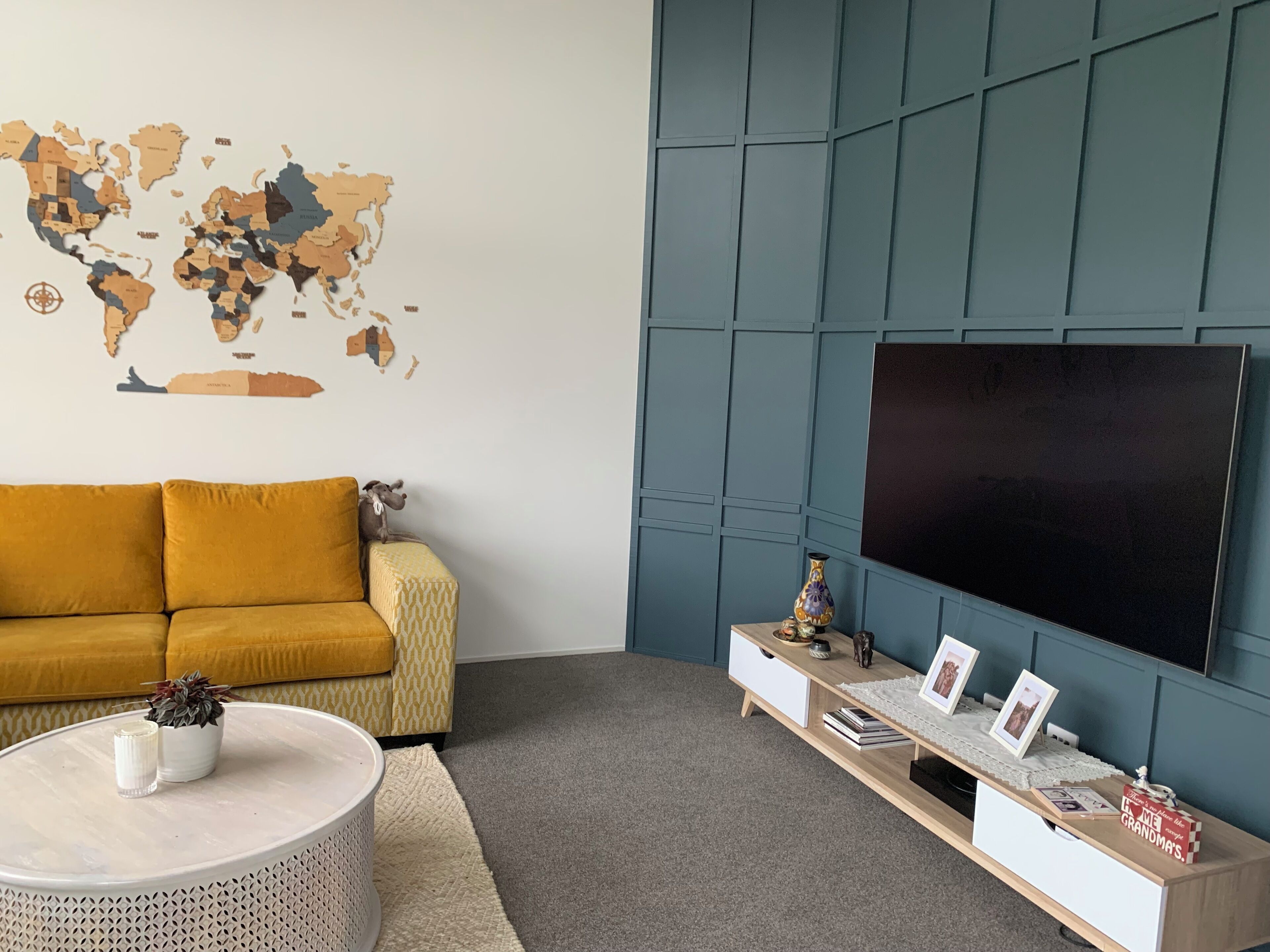 Un salon avec un canapé de couleur moutarde, une carte du monde accrochée au mur, un téléviseur à écran plat, une petite table basse ronde et une table d’appoint en bois agrémentée d’objets décoratifs.