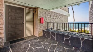 A stone-walled building with a closed door, a small red mailbox, and a balcony overlooking the ocean. - Direct Ocean Front Steps From the Beach Large Lanai - Vrkcv (Kailua-Kona)