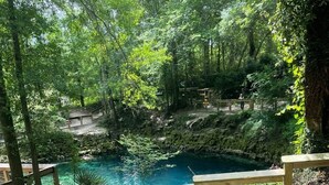 Une terrasse en bois mène à une piscine naturelle entourée d’arbres. Un panneau d’avertissement indique qu’il n’y a pas de sauveteur et que la baignade se fait à vos propres risques.