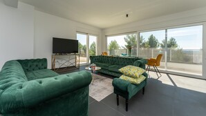 A modern living room with a green sofa set, a flat-screen TV on a stand, a dining table with chairs, and a balcony with a view of trees and a fence. - Luxurious home in Kakma (Kakma)