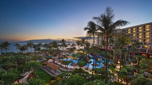 A resort with a large pool area, palm trees, and a view of the ocean at dusk.