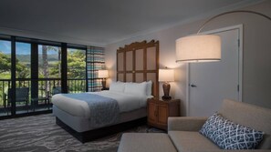 Une chambre d’hôtel avec un grand lit, une table de chevet avec des lampes, un canapé et une vue sur la verdure grâce à de grandes fenêtres.