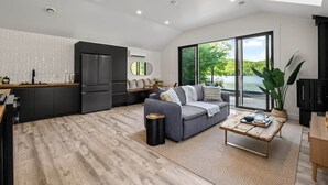 Un espacio habitable moderno con cocina, un sofá gris, una mesa de centro de madera y un amplio ventanal con vistas a un lago.