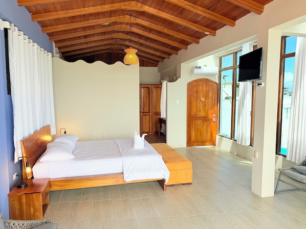 Un dormitorio con techo de madera, una cama con ropa blanca, una mesita de noche con lámpara y un televisor montado en la pared.