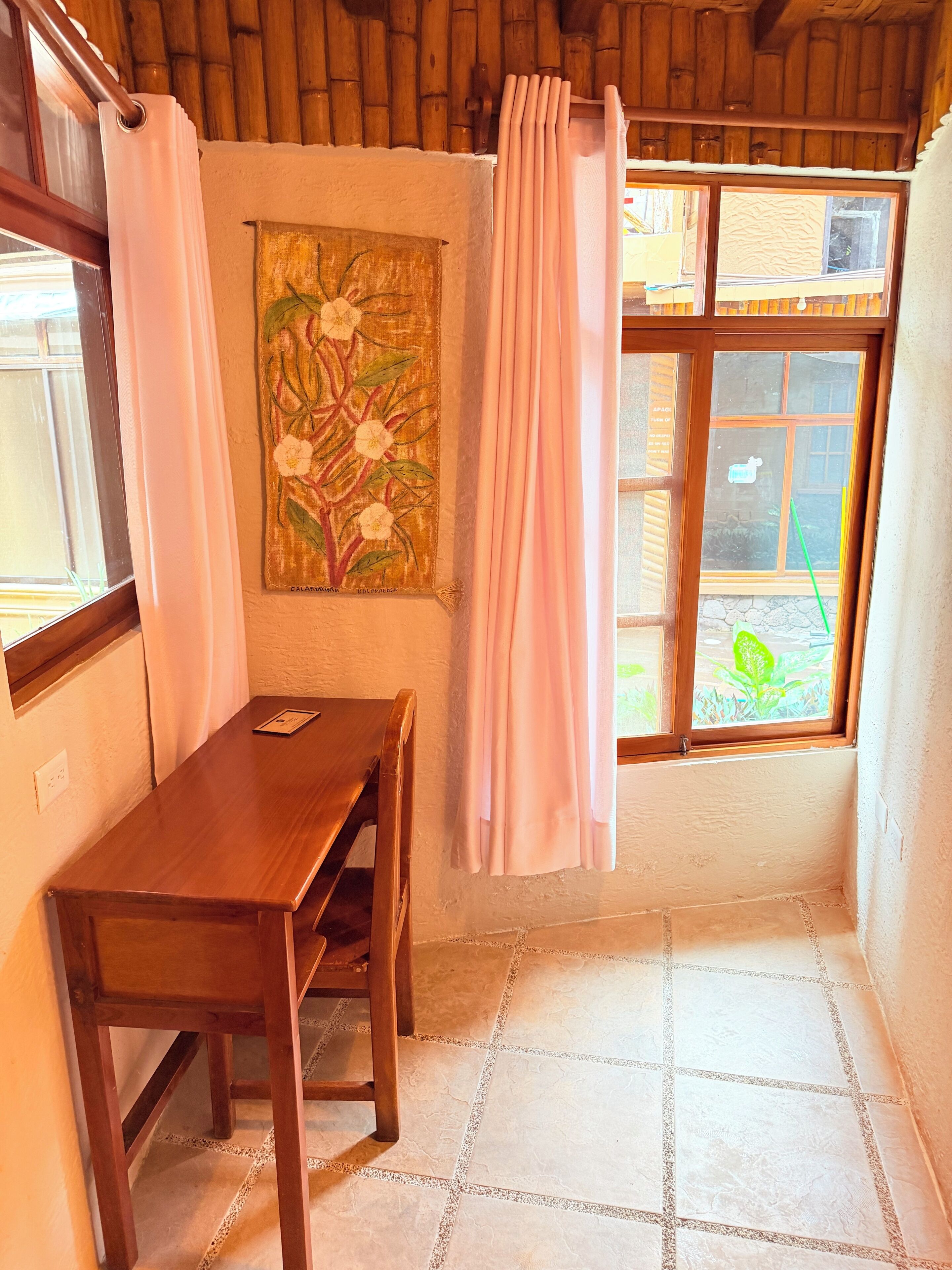 A corner room with a wooden table, a painting on the wall, a window with curtains, and a tiled floor.