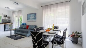 Un salon moderne avec un canapé gris, des chaises noires et blanches, une table à manger avec des assiettes et une bouteille, et un coin cuisine avec un réfrigérateur et une cuisinière.