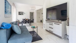 Un salon moderne avec un canapé gris, un coin repas avec des chaises noires et blanches, une cuisine dotée d’une grande télévision et une porte blanche donnant sur une autre pièce.