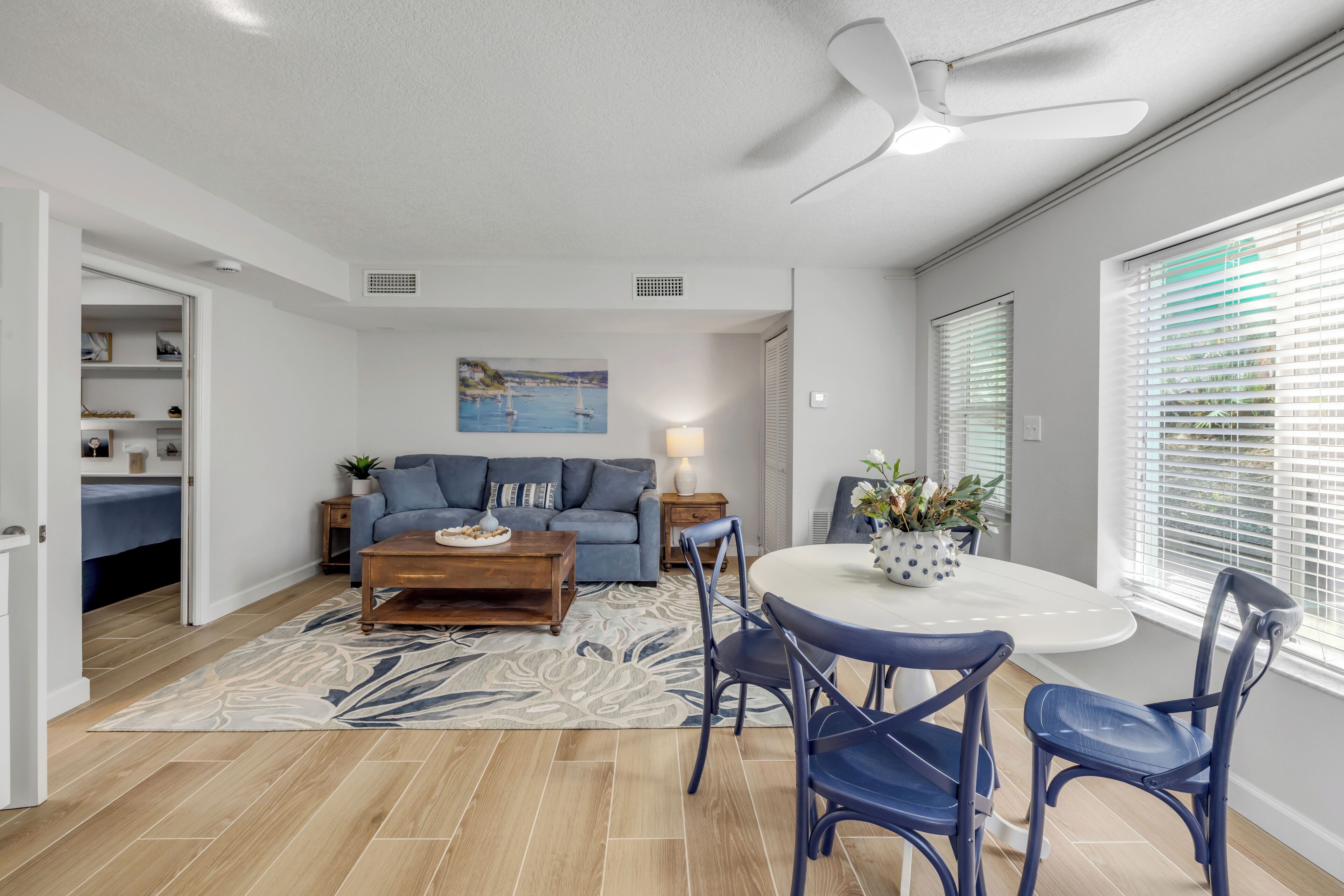 Un salon moderne avec un canapé, une table basse et un coin repas. On y trouve un ventilateur de plafond, un tableau au mur et une fenêtre avec des stores.