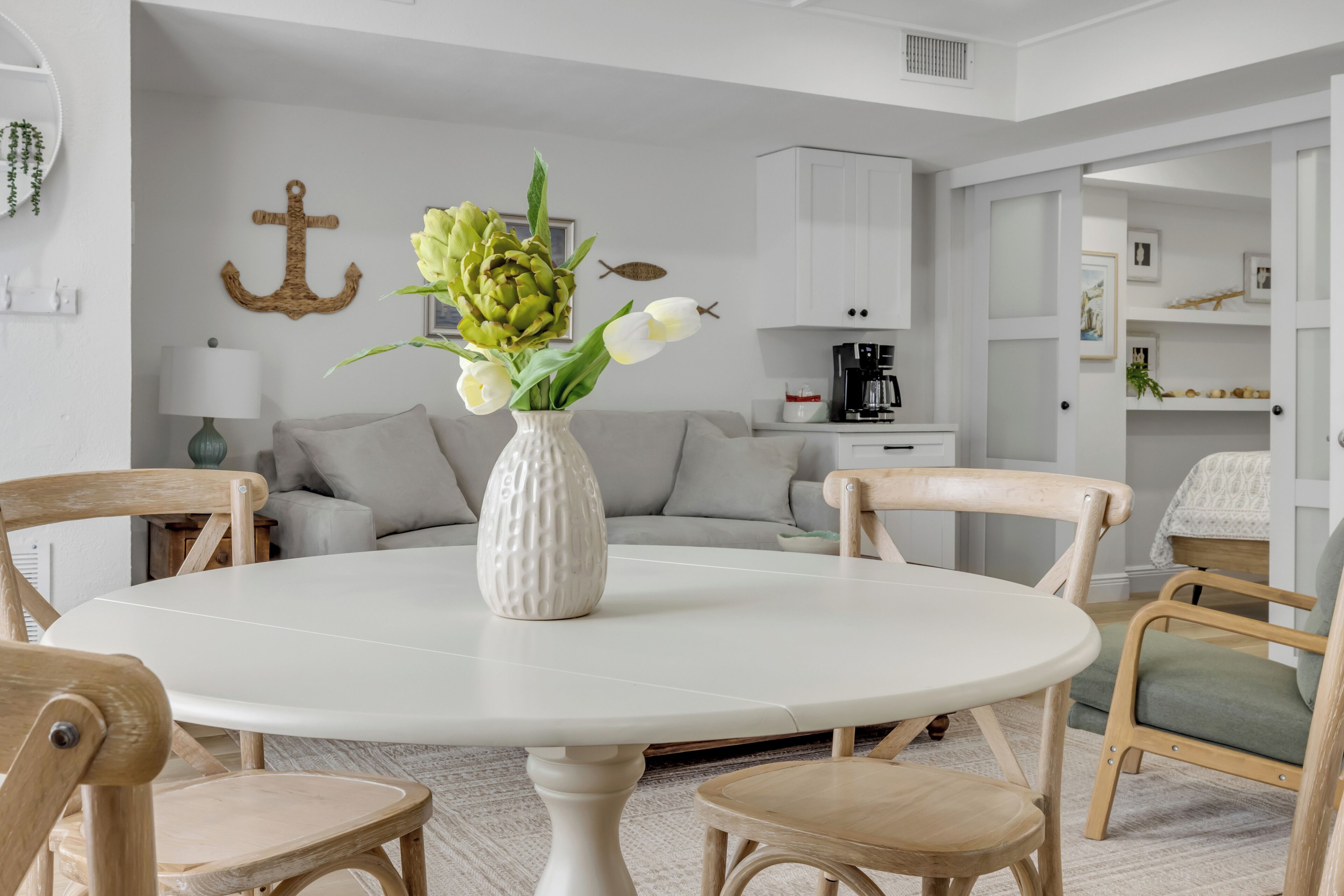 A dining area with a round table, chairs, a vase with flowers, a sofa, and a kitchen in the background.
