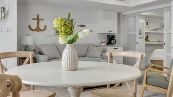 A dining area with a round table, chairs, a vase with flowers, a sofa, and a kitchen in the background.
