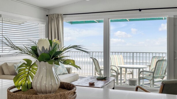 A balcony with a view of the ocean, a white sofa, a wicker table, and striped chairs.