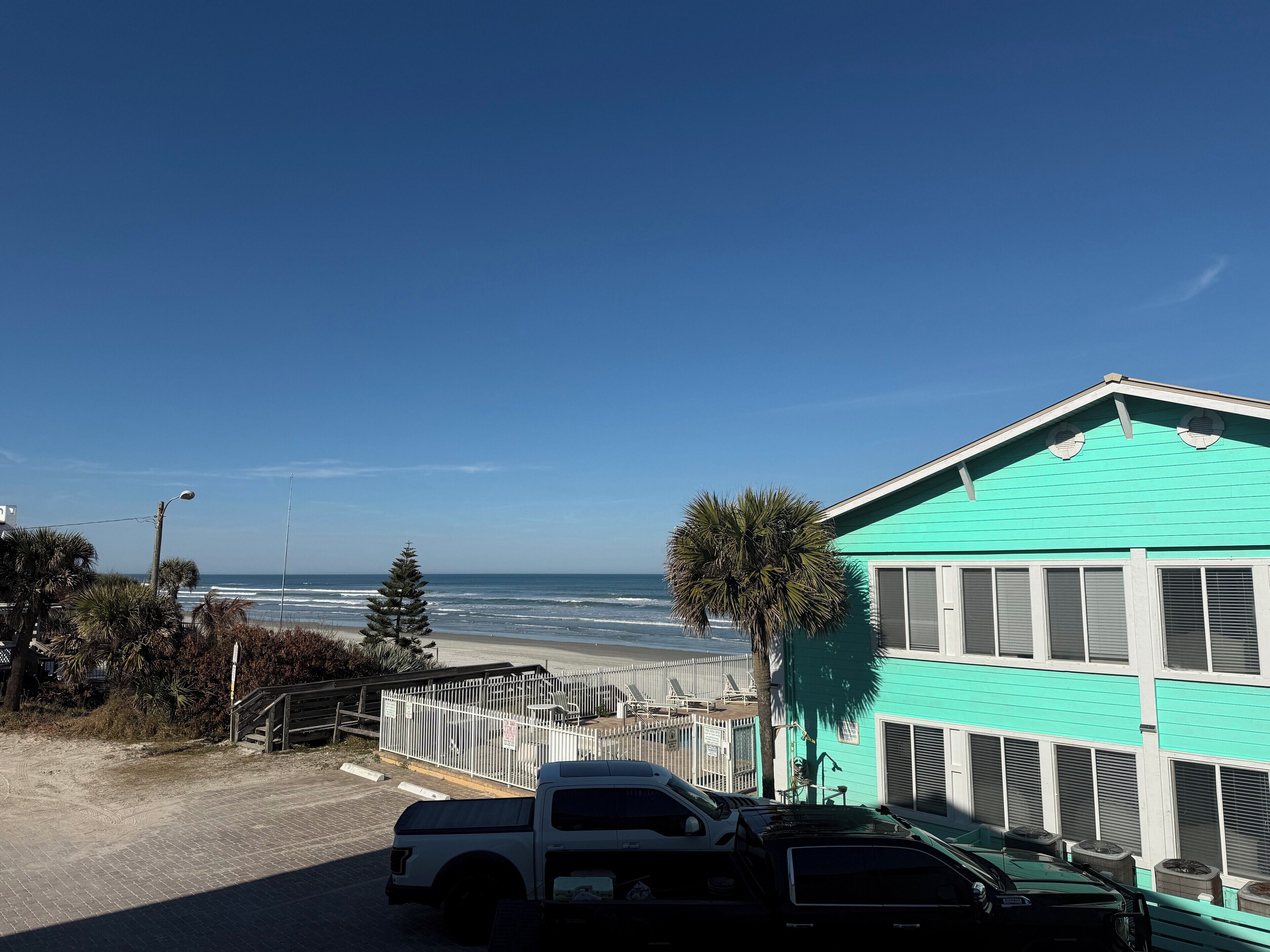 Un bâtiment turquoise avec vue sur la plage, un espace de stationnement avec un camion blanc et un chemin piétonnier délimité par une clôture.