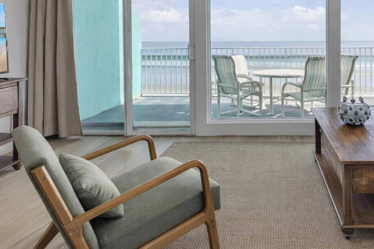 Sala de estar com sofá, mesa de centro de madeira e uma cadeira. Há uma televisão em um suporte à esquerda. O quarto tem uma grande porta de vidro deslizante que dá para uma varanda com vista para a praia e o oceano.