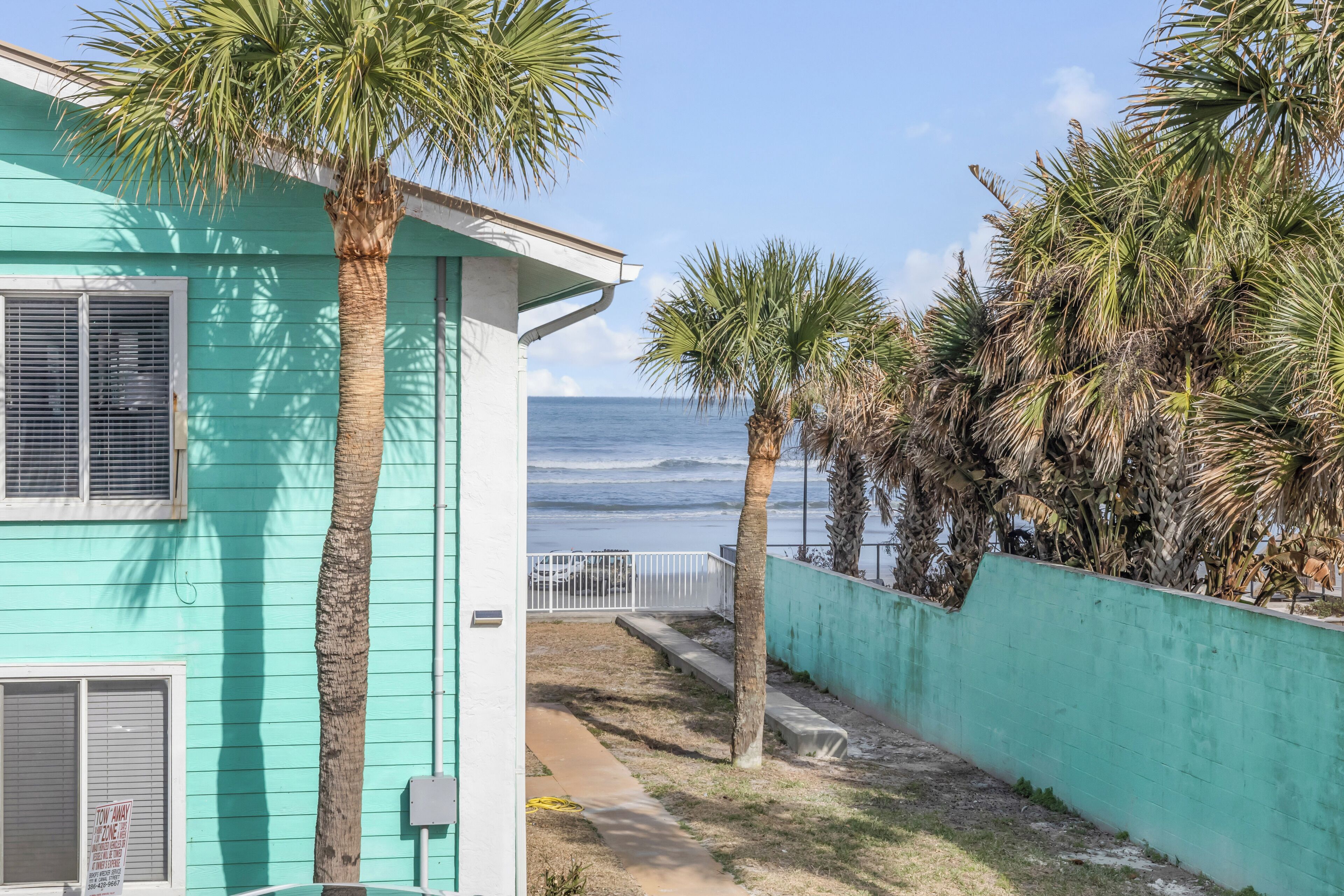 A turquoise building with a view of the ocean, palm trees, and a fenced pathway.