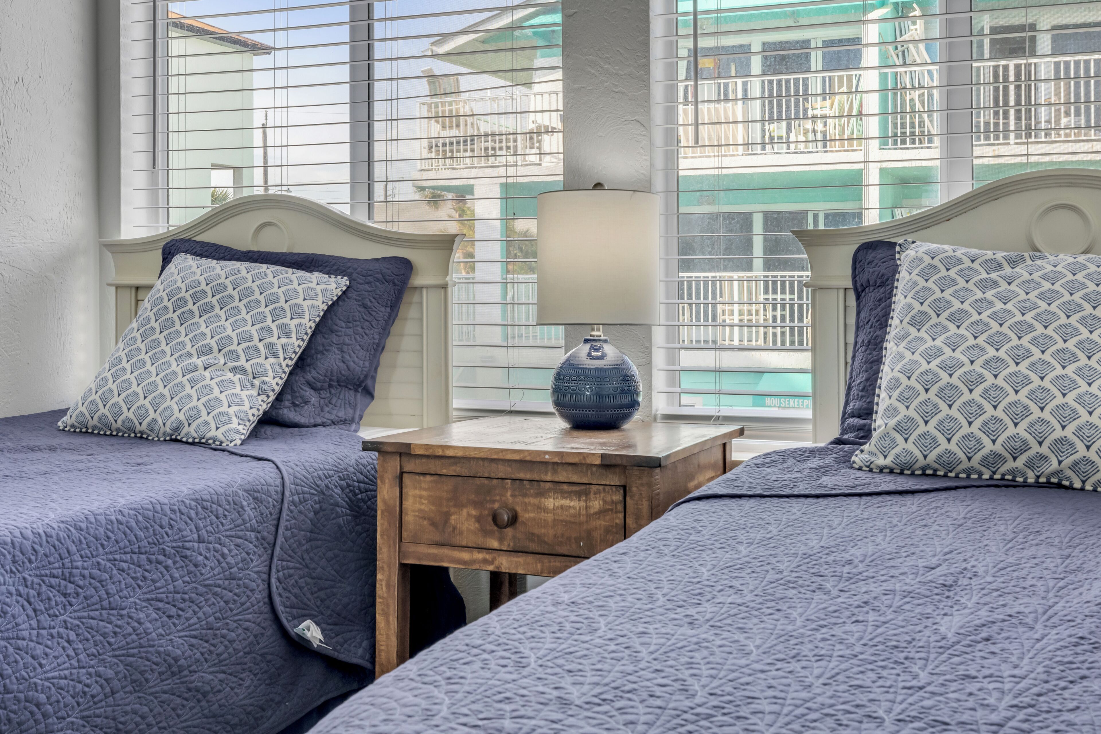A bedroom with two beds, a nightstand, a lamp, and a window with a view of buildings.