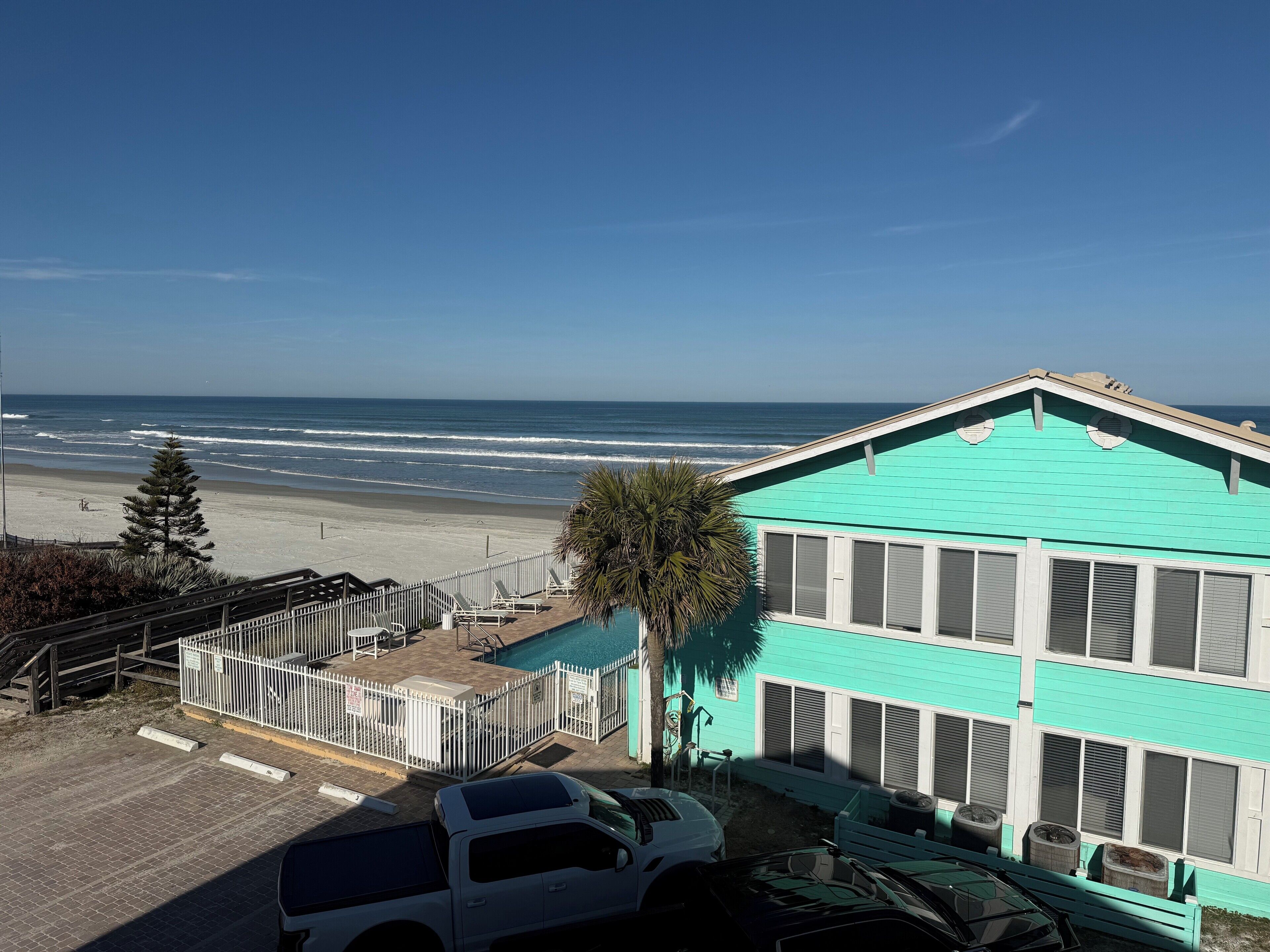 A turquoise building with a pool, a beach view, and a parking area with cars.