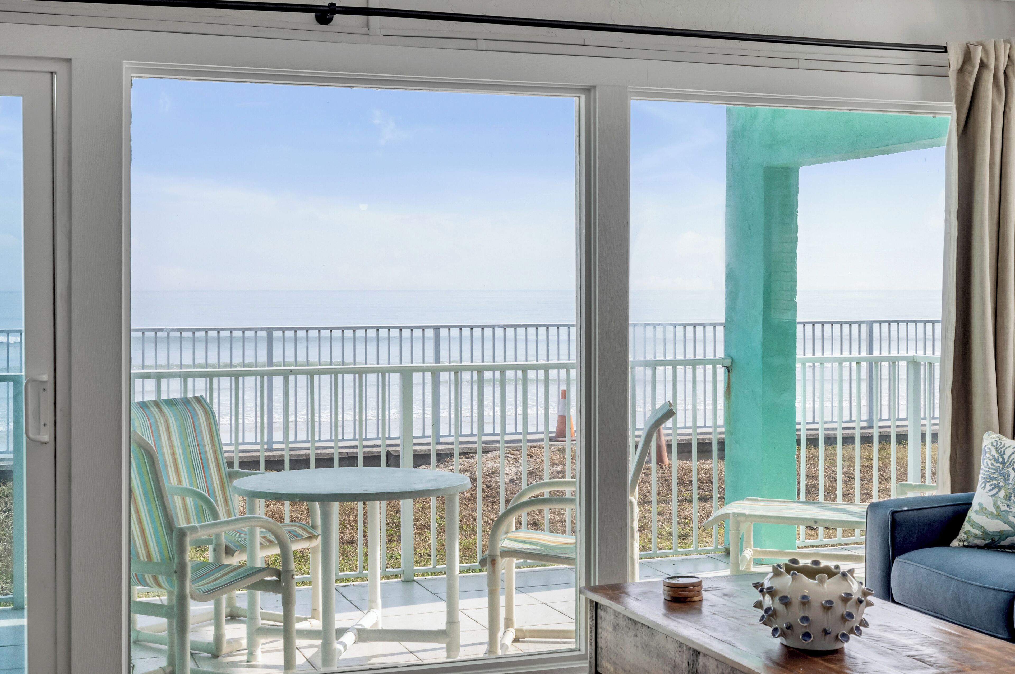Een balkon met uitzicht op zee, witte stoelen, een kleine tafel en een blauwe bank met een decoratieve vaas.