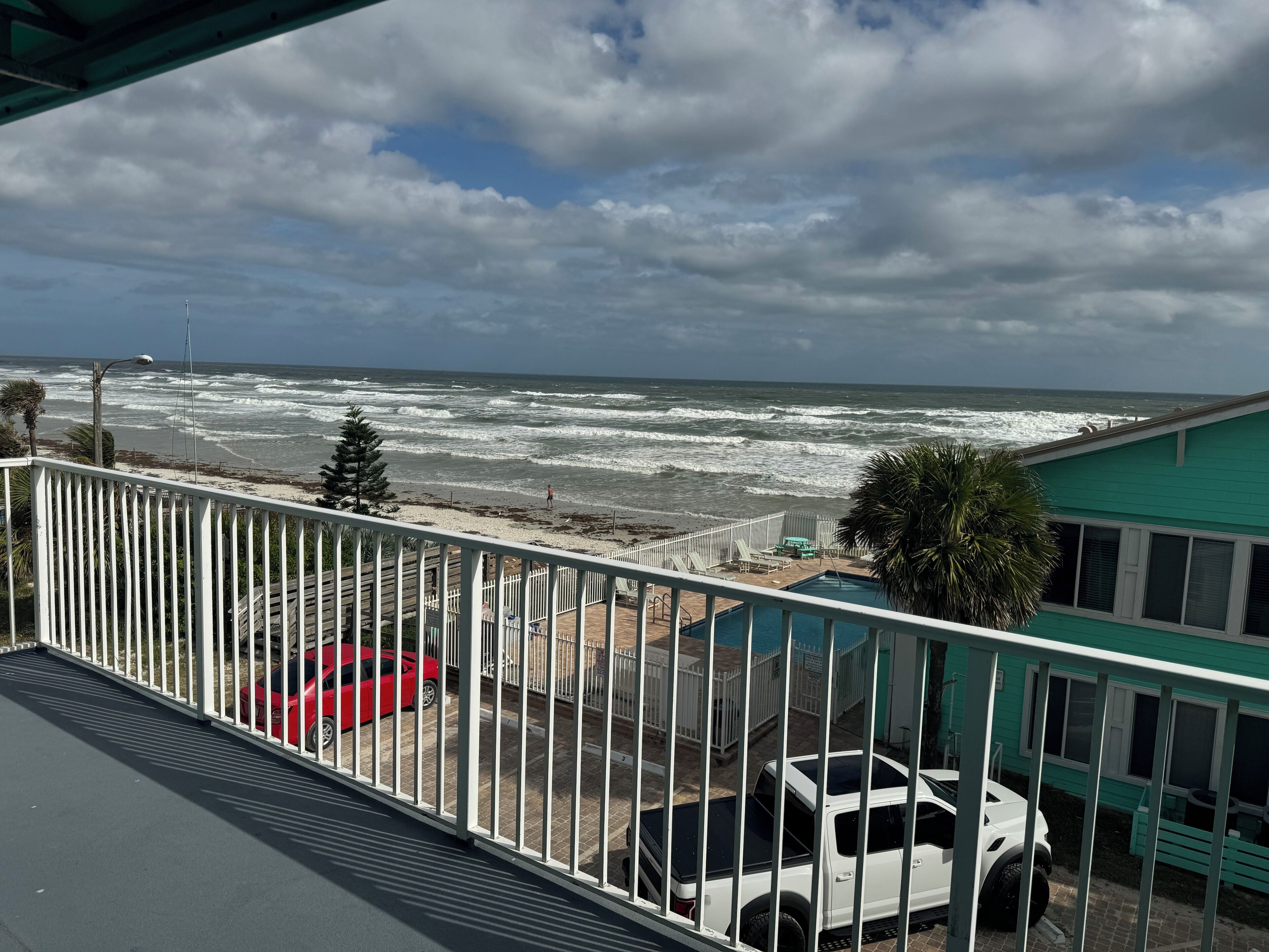 A balcony with a view of the ocean, a white fence, and parked cars.
