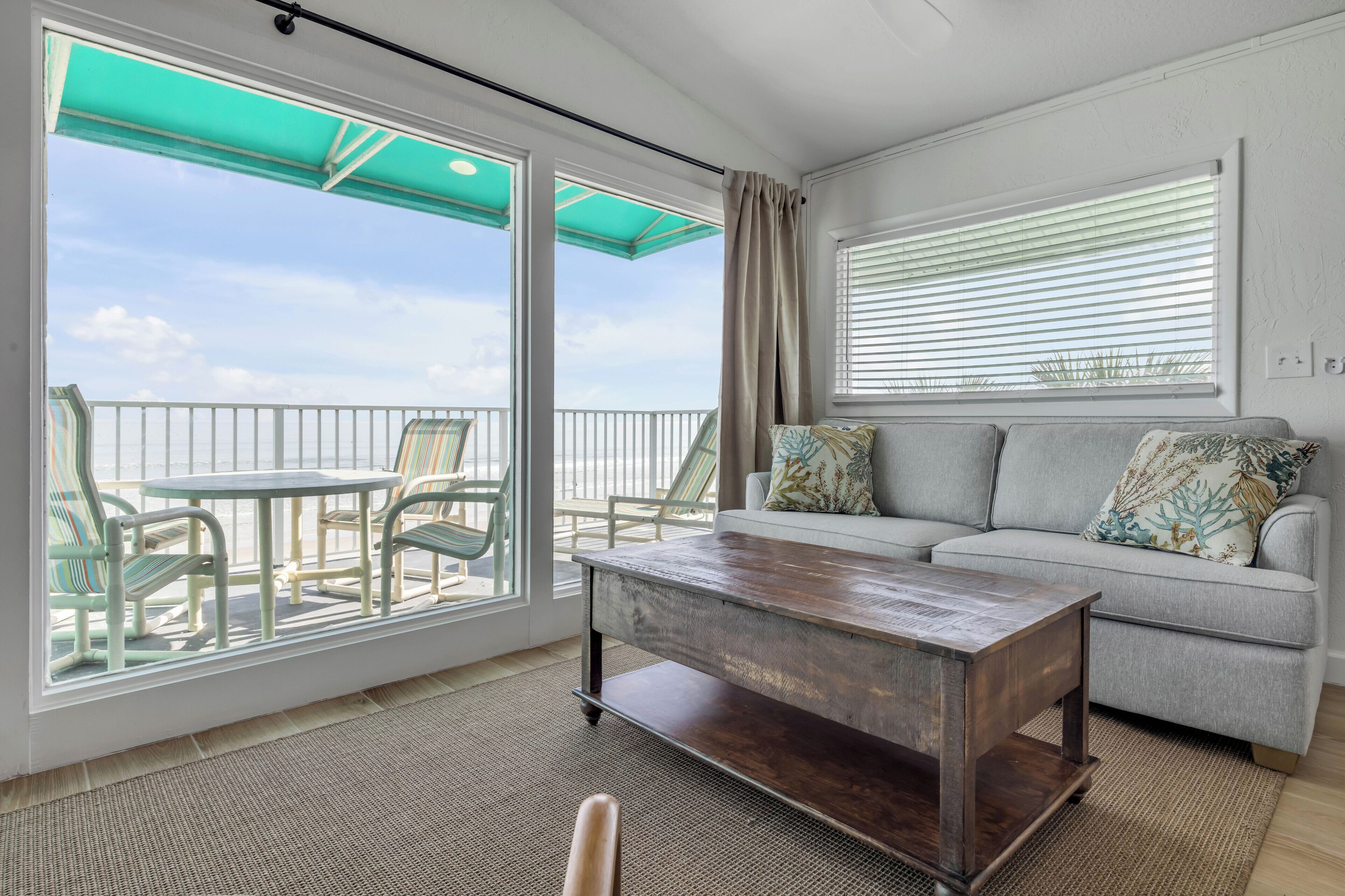 Ein Balkon mit Blick auf den Ozean, ein graues Sofa mit gemusterten Kissen und ein Holztisch.