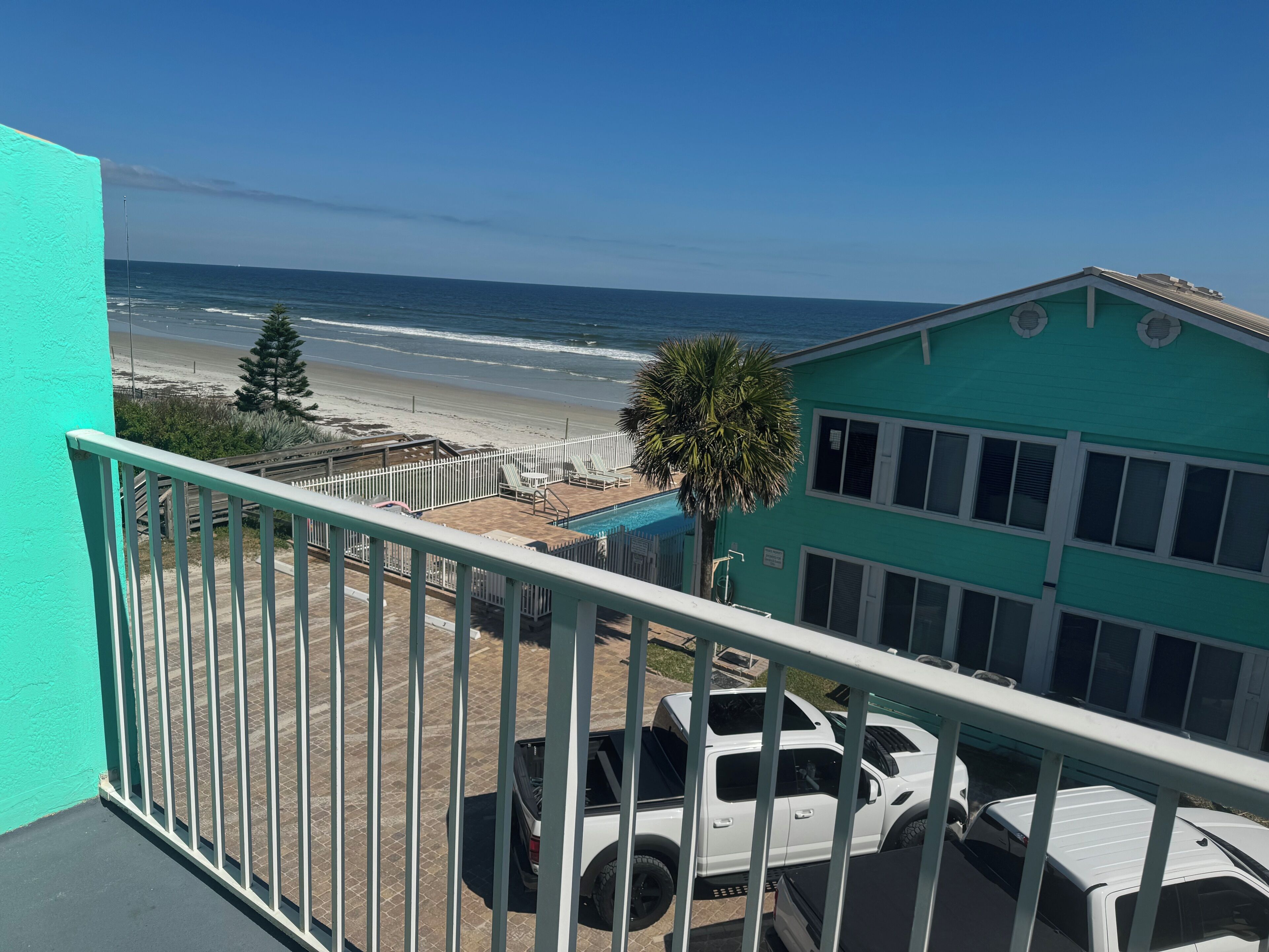 A balcony with a view of the beach and ocean, a white pickup truck, and a turquoise building.
