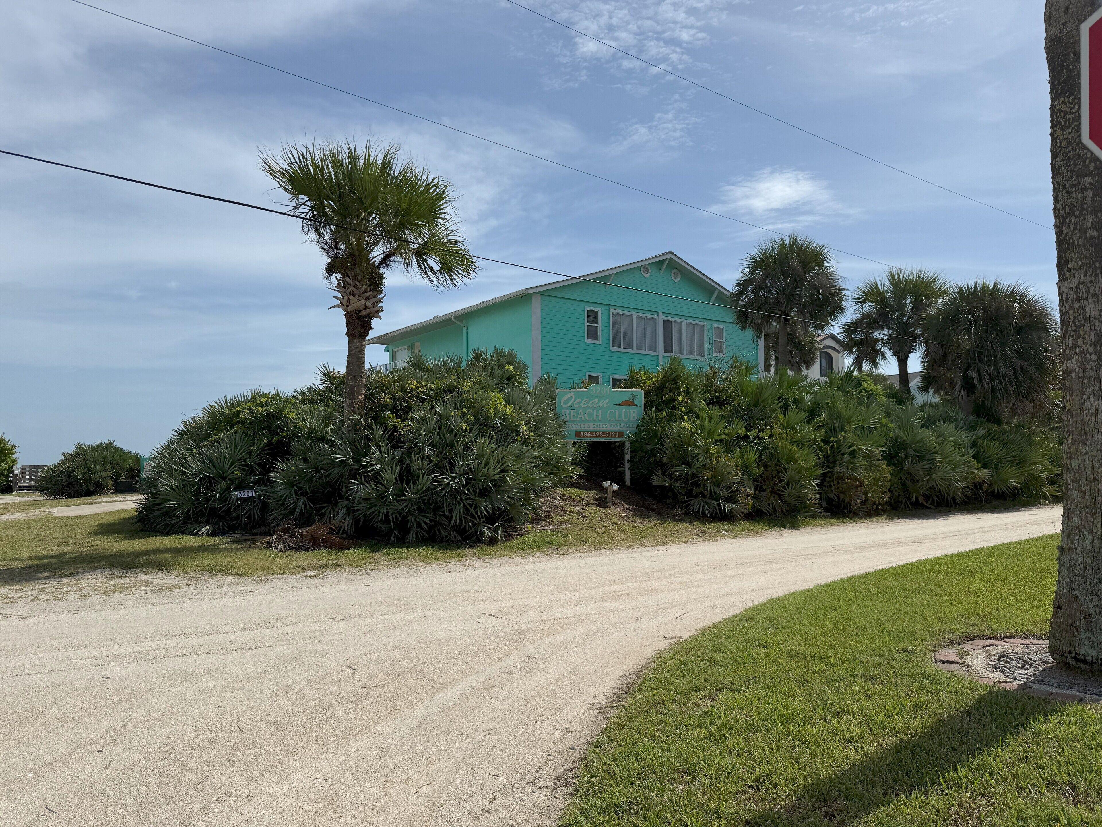 A turquoise building with a sign for Ocean Beach Club, surrounded by lush greenery and a dirt road.