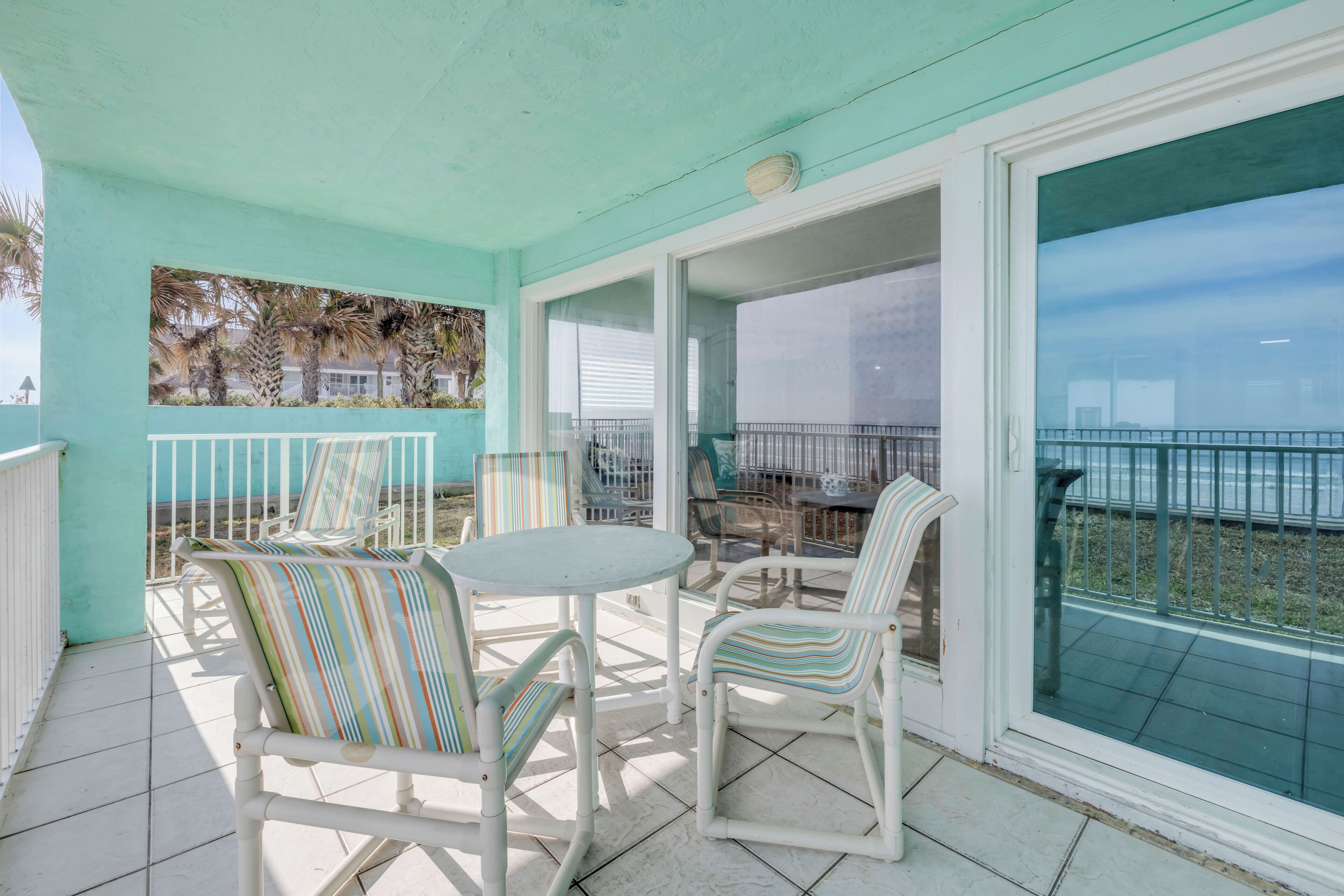 A covered outdoor area with white chairs and a table, a view of the sea through large glass doors, and a turquoise wall.