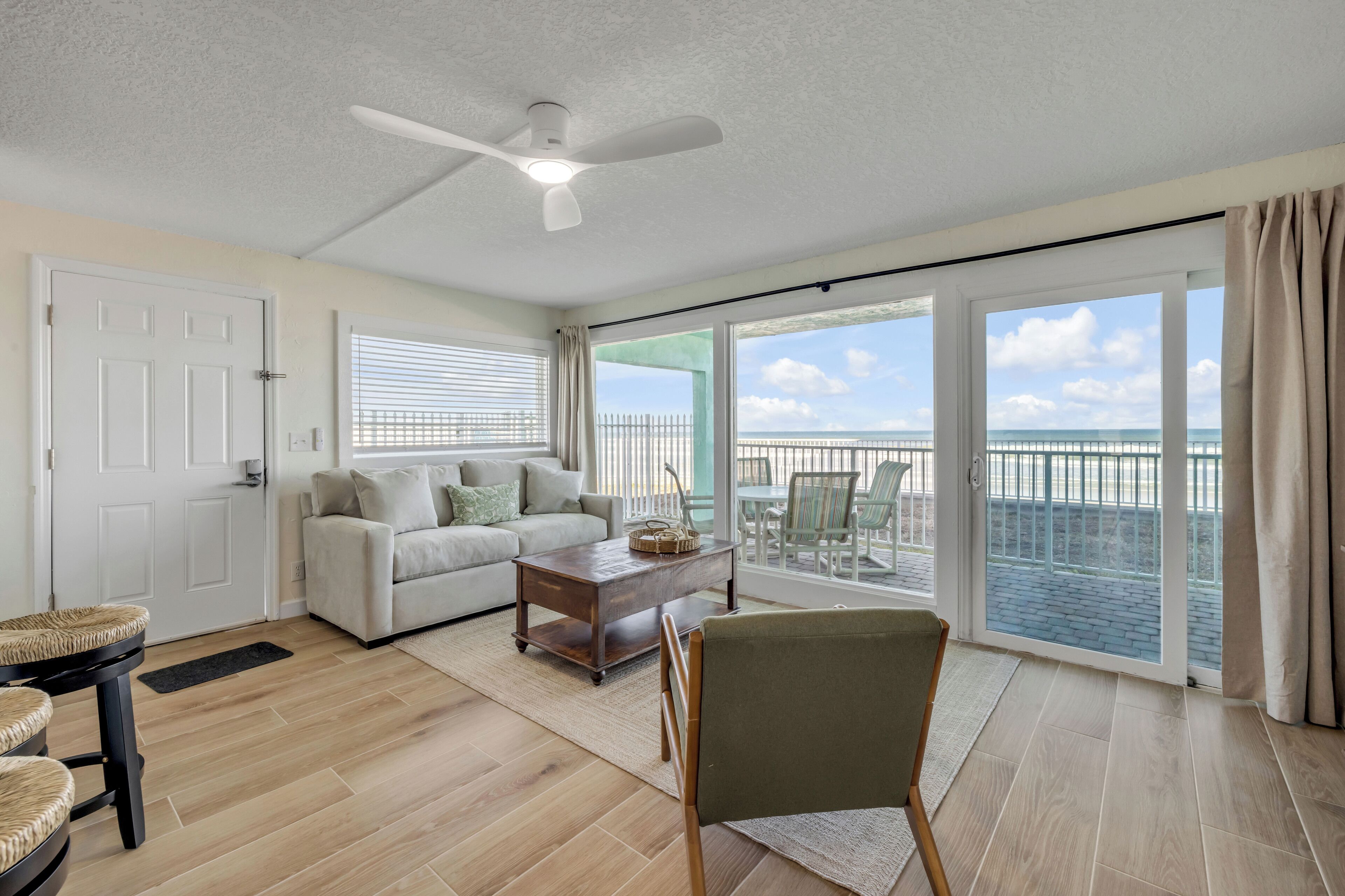 A spacious living room with a sofa, coffee table, and a chair. A ceiling fan is visible, and there is a large sliding glass door leading to a balcony with a view of the ocean.