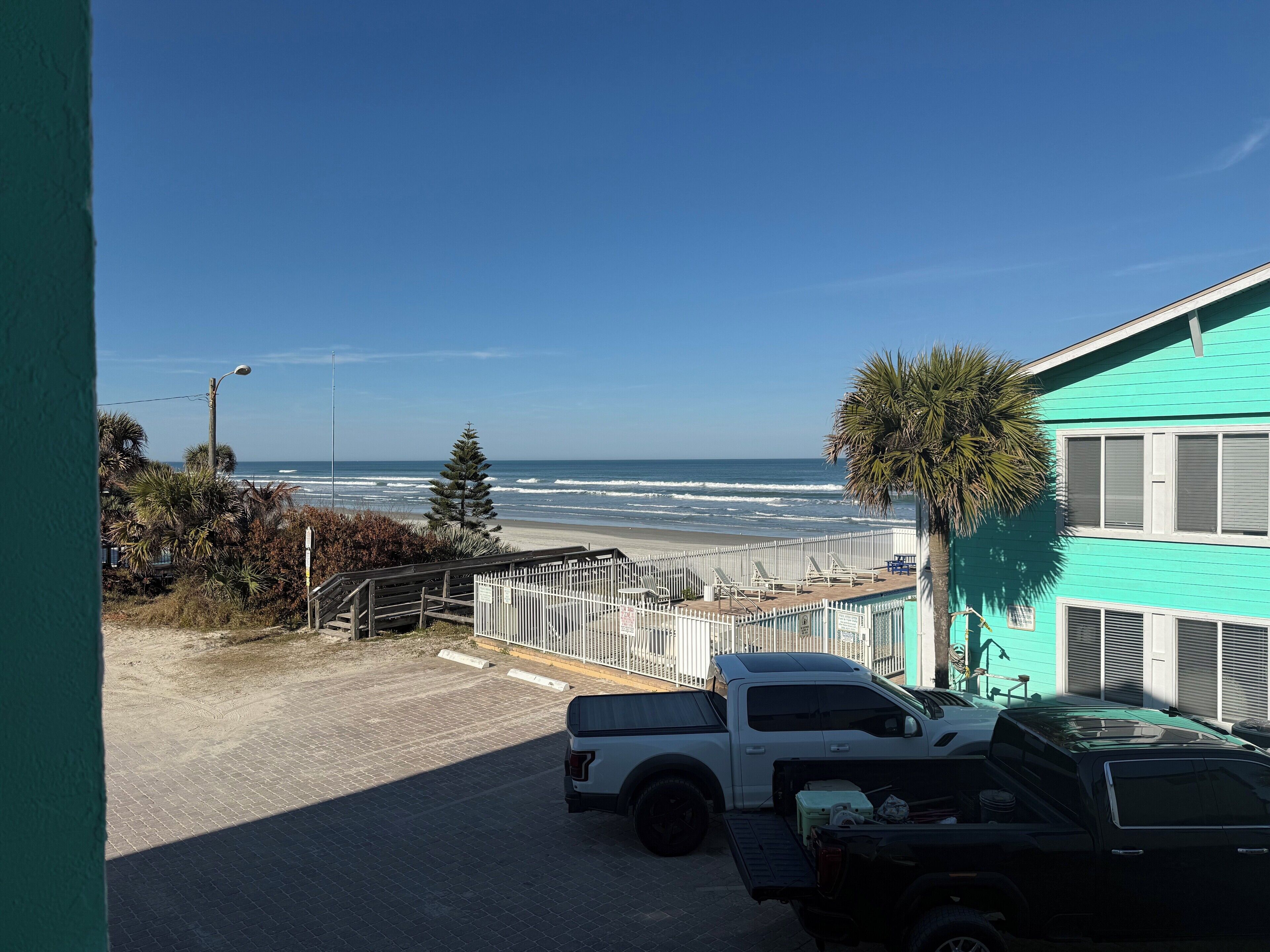 A view of a beach with a white pickup truck parked in a driveway, a turquoise building, and a clear blue sky.