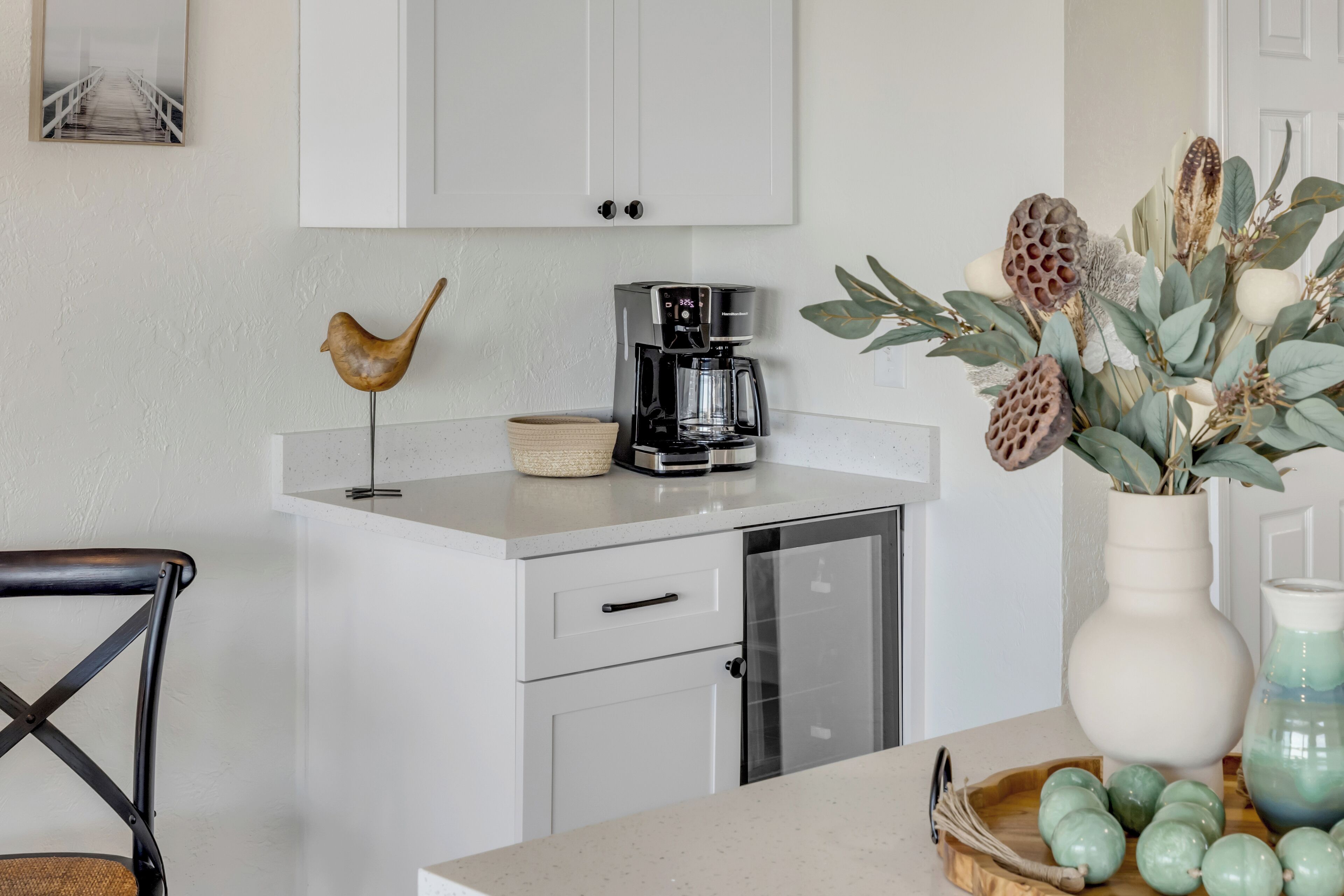 A kitchen with white cabinets, a coffee maker, a decorative bird, a vase with green apples, and a wicker basket.