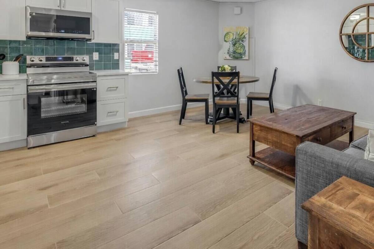 Cozinha moderna com armários brancos, forno embutido e micro-ondas. Há uma sala de jantar com mesa e cadeiras de madeira. Um sofá cinza e uma mesa de centro de madeira estão na sala de estar.