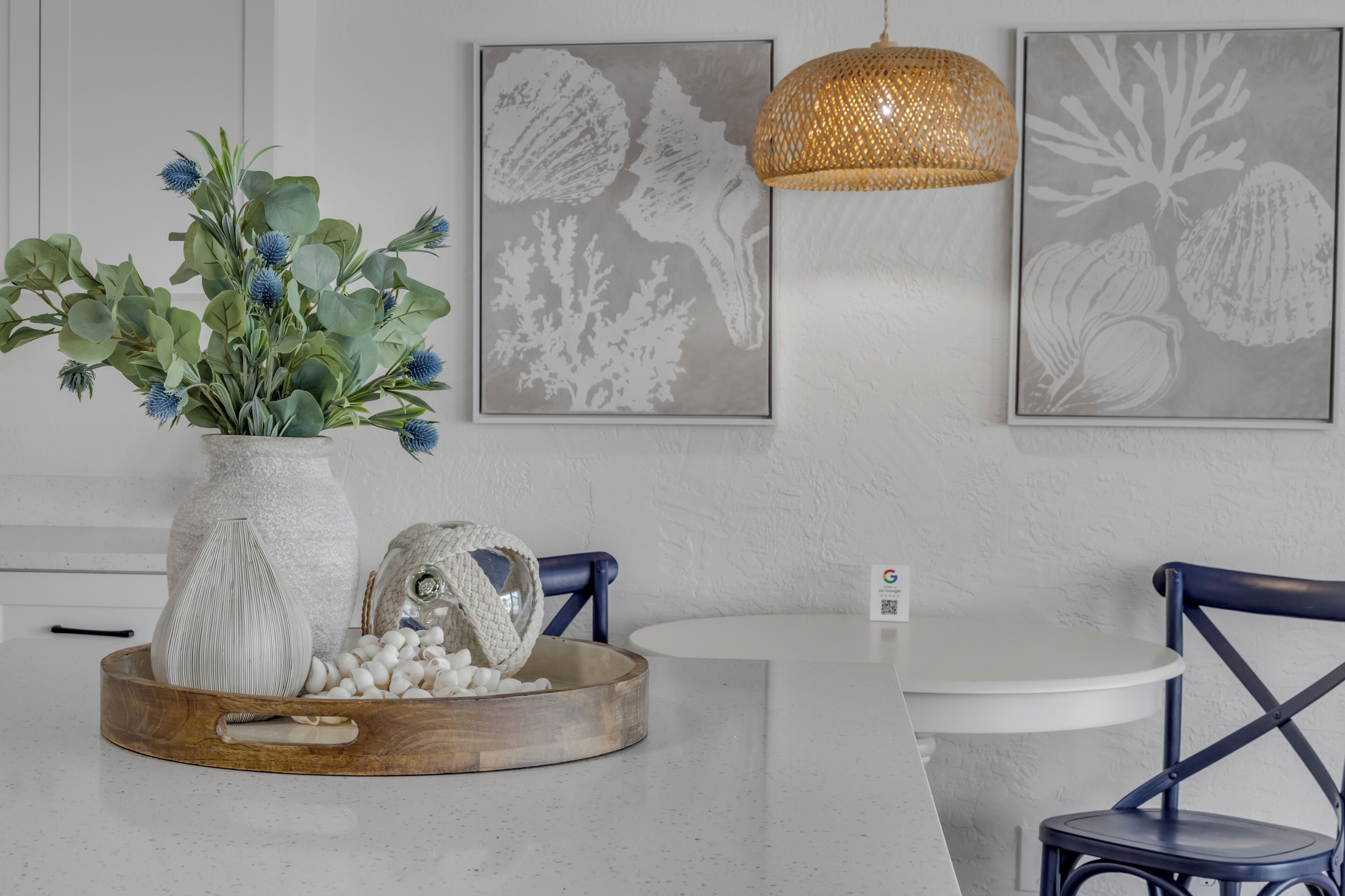 Un comedor con una mesa blanca, una silla azul, un jarrón con plantas verdes y dos obras de arte enmarcadas en la pared.