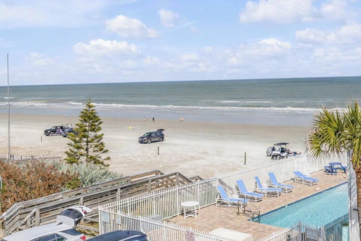 Vista da praia com piscina, espreguiçadeiras, carrinho de golfe e um prédio turquesa.