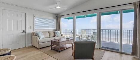 A spacious living area with a sofa, coffee table, and a chair, a balcony with a view of the sea, and a ceiling fan.
